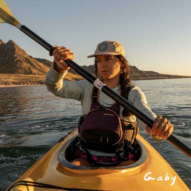 A woman paddling in a yellow kayak on the water at sunset, wearing a patterned cap, light long-sleeve top, and a dark purple and black kayak backpack. The word 'Gaby' is written in white script in the bottom right corner.
