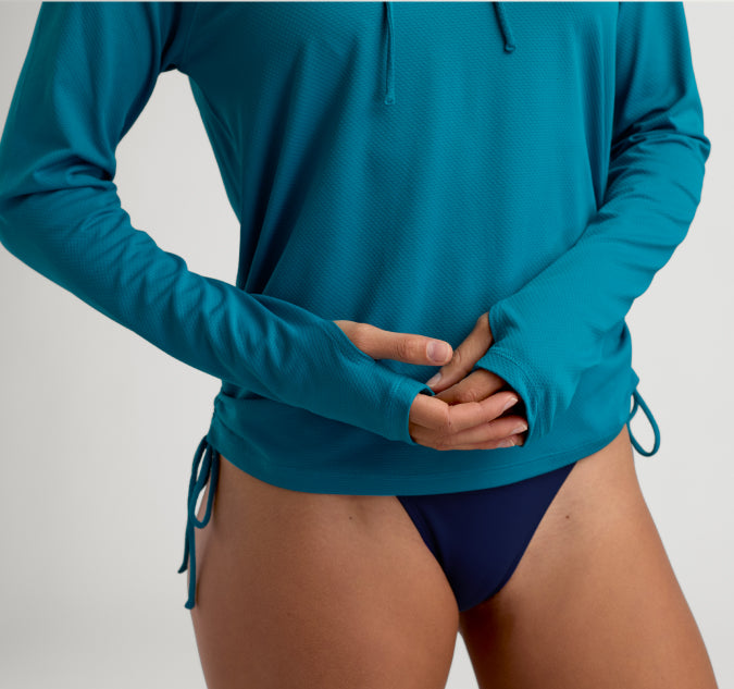 A close-up of a model wearing a teal long-sleeve swim shirt with thumbholes and side ties, paired with dark navy bikini bottoms, against a light gray studio background.