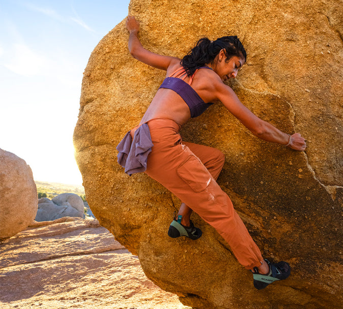 A woman boulders on a large tan rock in a desert setting, wearing an orange-brown crop top, a purple sports bra, and black climbing shoes.