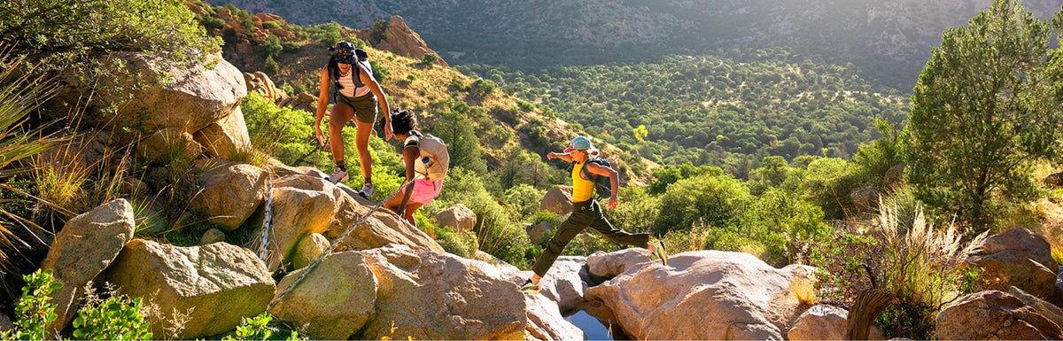 Three people hiking up a rocky, grassy mountain slope in a sunny outdoor setting with green forested hills in the background.