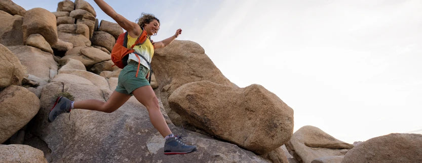 A woman hiking up a rocky mountain slope, wearing a yellow and green athletic top, green shorts, and hiking boots, with an orange backpack against a bright sky.