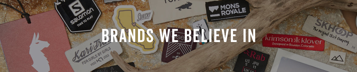 A collage of various embroidered patches displayed on a rustic wood background, featuring prominent brands such as Salomon, Rab, Sunski, KariTraa, Cotopaxi, and Skhoop. A bold white text overlay in the center reads 'BRANDS WE BELIEVE IN'.