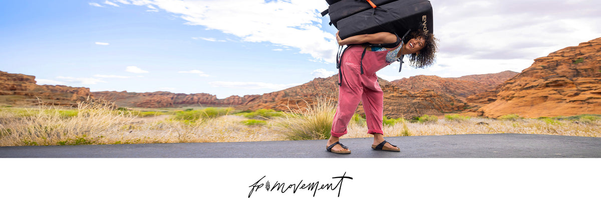 A person outdoors in a desert landscape, carrying a large black duffel bag on their back while wearing pink pants and sandals. The photo has a wide panoramic composition with a white band at the bottom containing the brand name 'fp movement'.