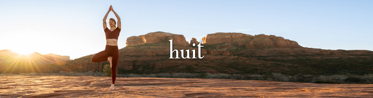 A woman in a yoga pose stands outdoors in front of rocky desert mountains at sunrise, with the Huit logo centered over the scene.