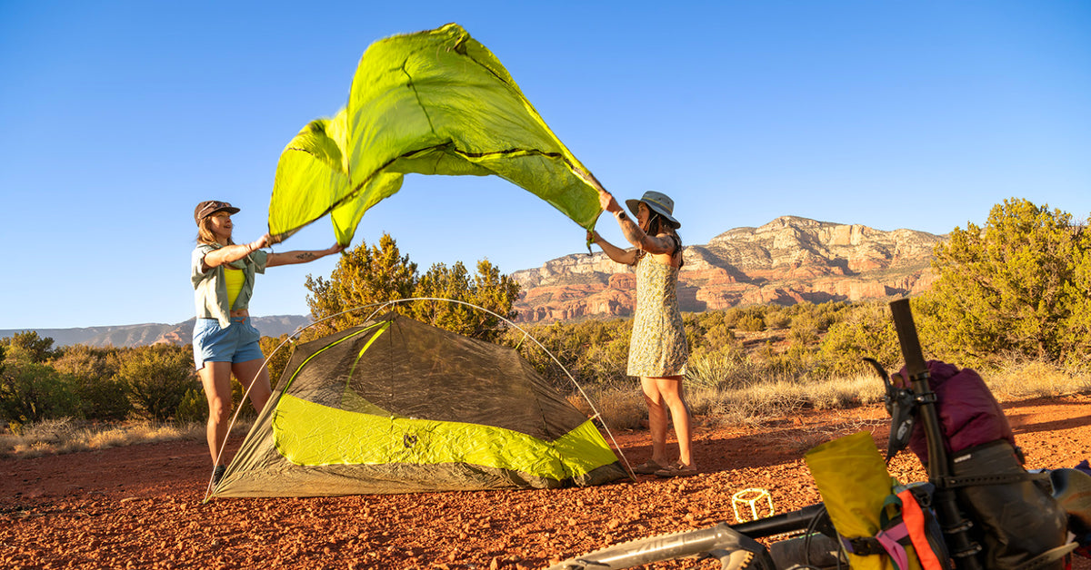 Two people assembling a bright lime green dome tent in an outdoor desert campsite with red dirt ground, shrubs, and rocky mountains under a clear blue sky.