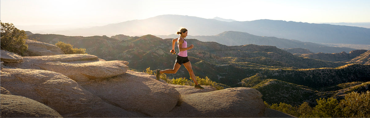 A woman running across large rock formations in an outdoor mountain landscape at sunrise or sunset, wearing a pink top and dark shorts.