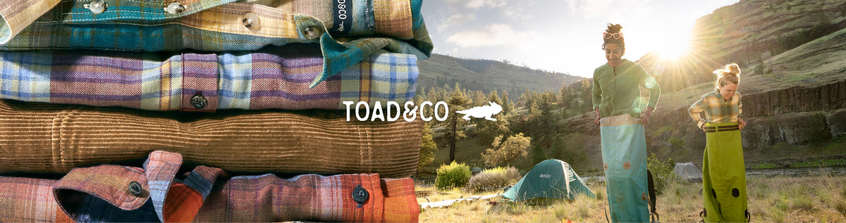 A stacked display of folded plaid and patterned camp shirts in various colors, with a scenic outdoor mountain camping lifestyle shot in the background featuring two models and a tent. The brand name 'TOAD&CO' is displayed in white text in the center.