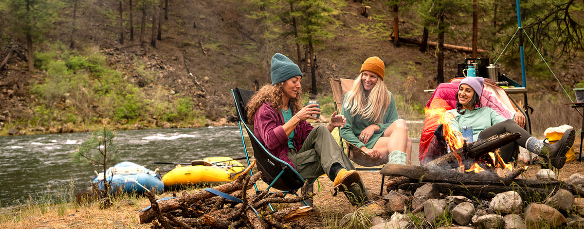 Three people relax in camping chairs by a river campsite, with a fire pit, logs, trees, and inflatable rafts visible in the outdoor setting.
