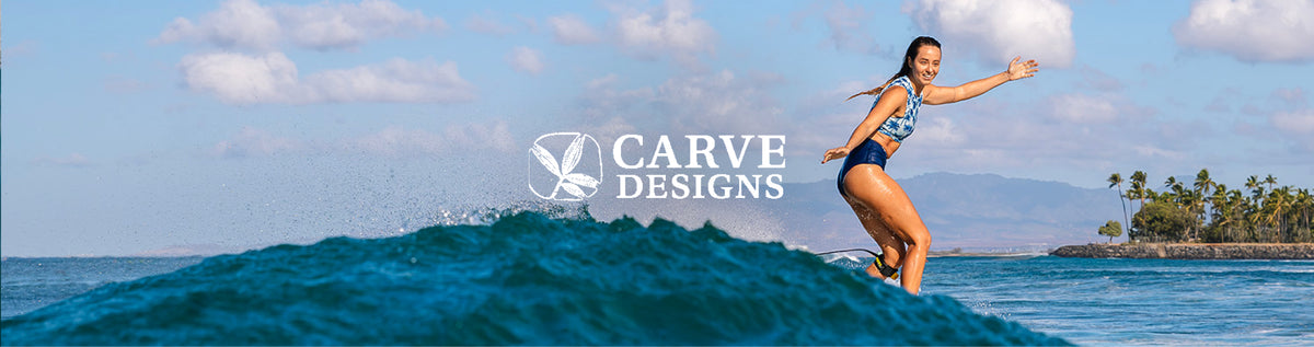 A woman in a blue patterned bikini stands on a surfboard on a turquoise ocean wave under a blue sky with clouds, with the text 'CARVE DESIGNS' overlaid in the center.