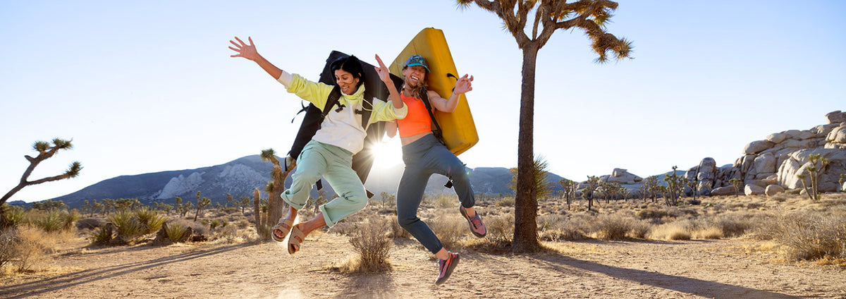 Two people jumping in a desert landscape under bright sunlight, carrying hiking gear and a yellow surfboard or paddle. A Joshua tree and rocky desert formations are visible in the background.