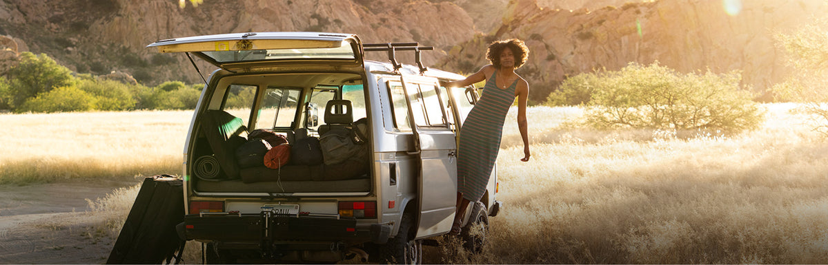 A woman in a sleeveless dress stands beside the open rear of a white camper van loaded with gear, parked in an open grassy landscape at golden hour.