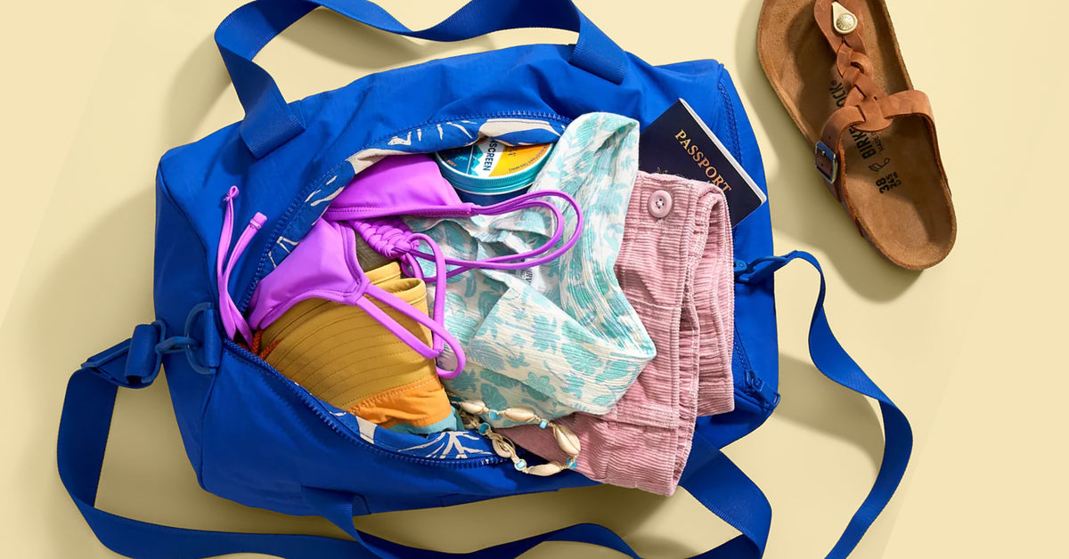 A bright blue duffel bag filled with colorful travel items, including a purple bikini top, patterned swim trunks, a yellow pillow, and a passport, shown on a beige background with a brown sandal nearby.