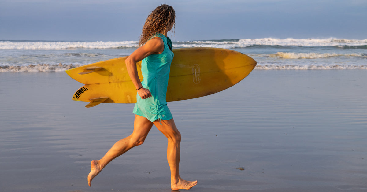 A woman carrying a yellow surfboard walks along the wet shoreline of a beach, wearing a turquoise sleeveless top and matching shorts.