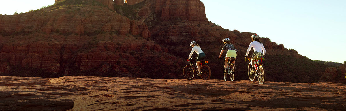 Three mountain bikers riding away from the camera on a rocky desert trail with large red rock formations in the background under a clear sky.
