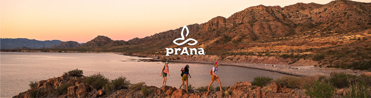 A scenic outdoor landscape at sunset with rocky cliffs, a shoreline, and water in the background. Three people are walking along the beach path, and a white 'prAna' logo is overlaid near the center.