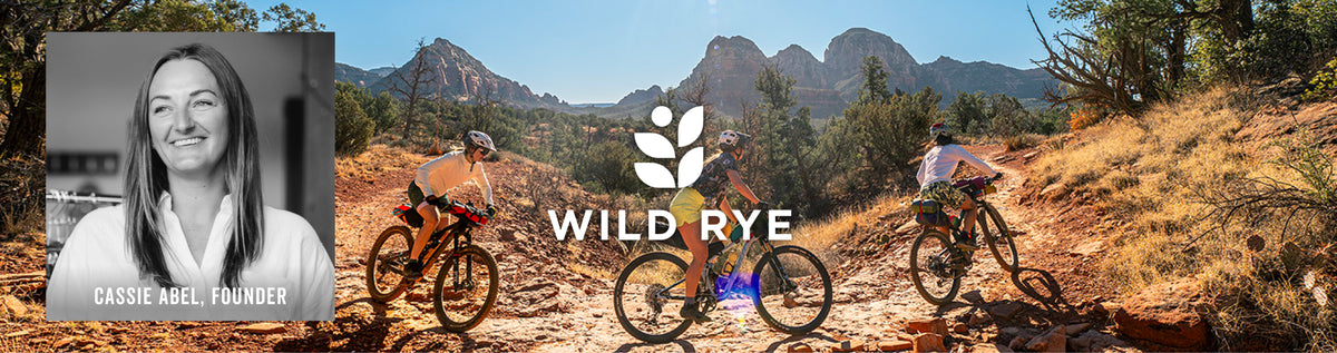 A promotional banner for Wild Rye featuring a black-and-white portrait of Cassidy Abel, founder, alongside three mountain biker action shots on a rocky desert trail with red rock mountains in the background. The Wild Rye logo is centered over the middle.