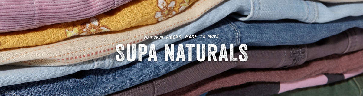A close-up stack of folded colorful garments or textiles with large white text reading 'SUPA NATURALS' across the center. The visible fabrics include pink, yellow, blue, beige, and brown tones, and the text appears to be a brand or collection name.