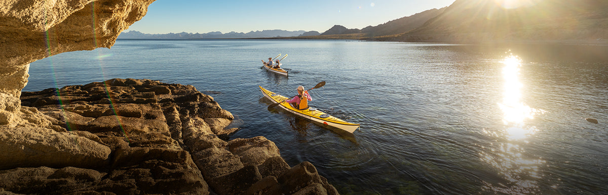 A scenic outdoor waterway at sunrise with two kayakers on calm blue water, framed by rocky cliffs in the foreground and distant mountains under a bright sunlit sky.