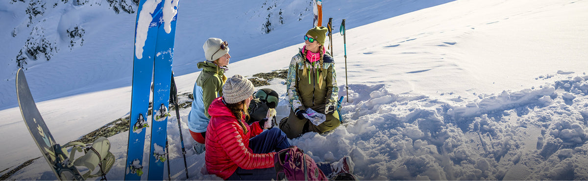 Three people sitting in deep snow with skis and ski poles nearby, dressed in winter outdoor gear against a snowy mountain background.