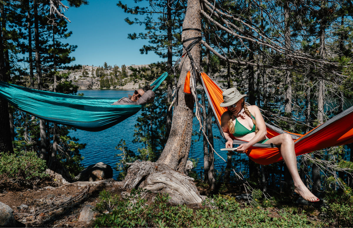 Two people relax in colorful hammocks strung between trees beside a blue lake, with forest scenery in the background. One hammock is teal and the other is orange-red.