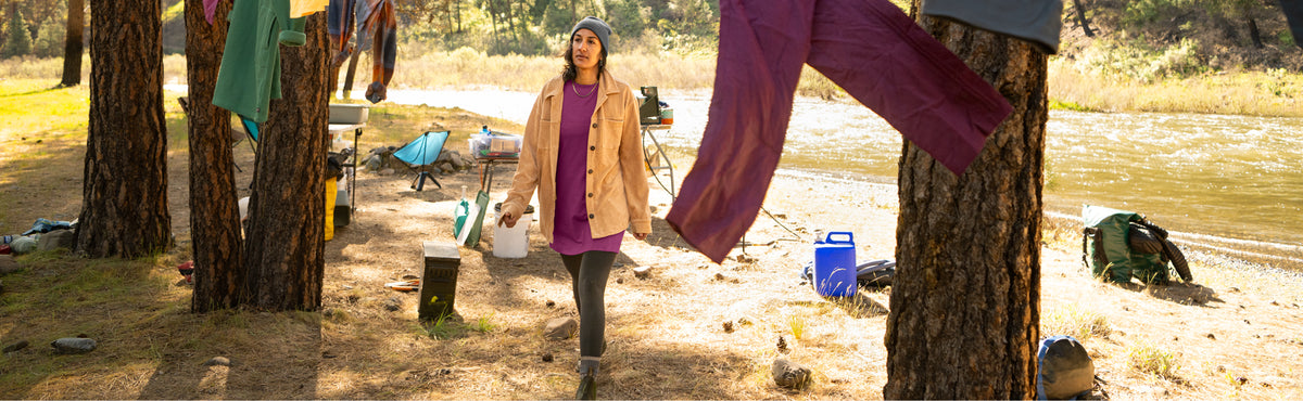 A woman stands at a lakeside campsite between trees, wearing a tan jacket over a purple top and dark leggings. Laundry hangs from trees nearby, and camping gear is scattered on the sandy shore.