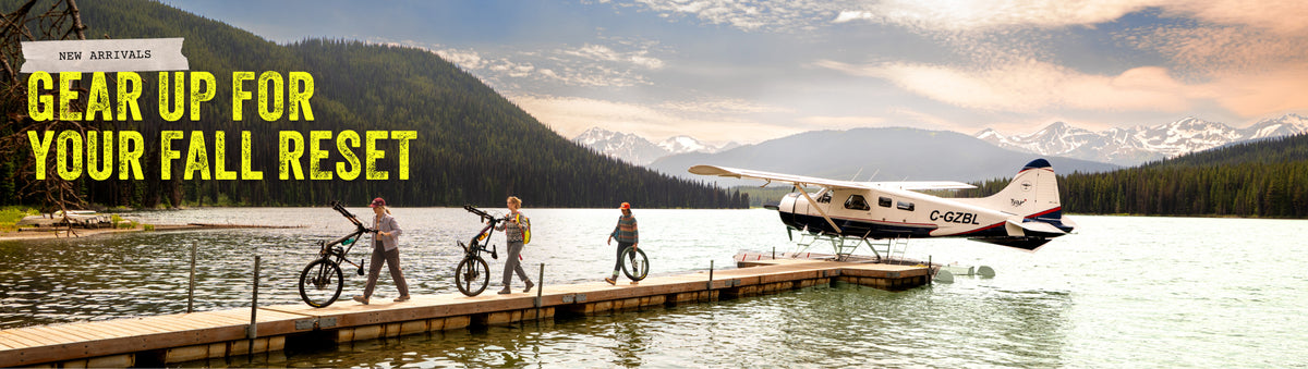 A scenic outdoor promotional banner showing a seaplane on a lake with four people walking along a dock carrying bicycles, set against mountains and trees. Large yellow text reads 'GEAR UP FOR YOUR FALL RESET' and a small badge says 'NEW ARRIVALS'.