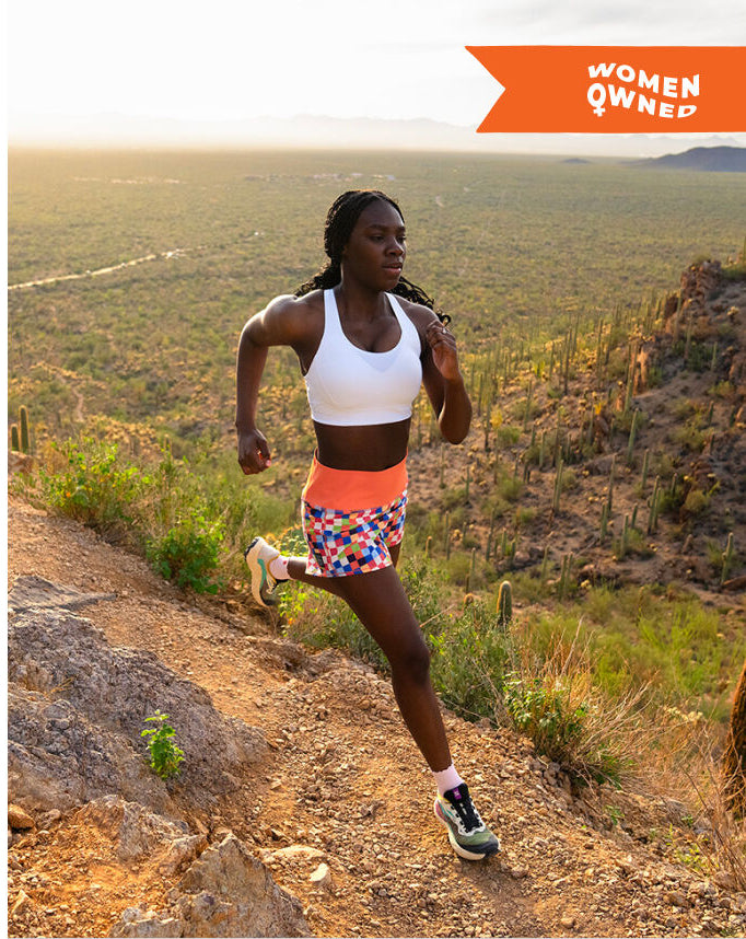 A woman running on a desert trail wearing a white sports bra and a colorful patterned athletic skirt, with a bright orange banner in the top right that reads 'WOMEN OWNED'.