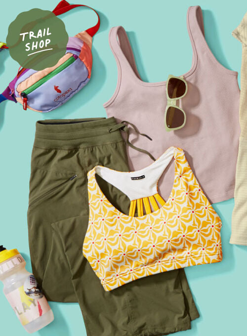 A flat lay of multiple women's activewear items and accessories on a light blue background, including a pale pink tank top, a cream zip-front athletic top, an orange sports bra, a yellow patterned sports bra, olive green shorts, yellow shorts, sneakers, sunglasses, a fanny pack, a cap, and a water bottle. A green circular badge in the top left reads 'TRAIL SHOP'.