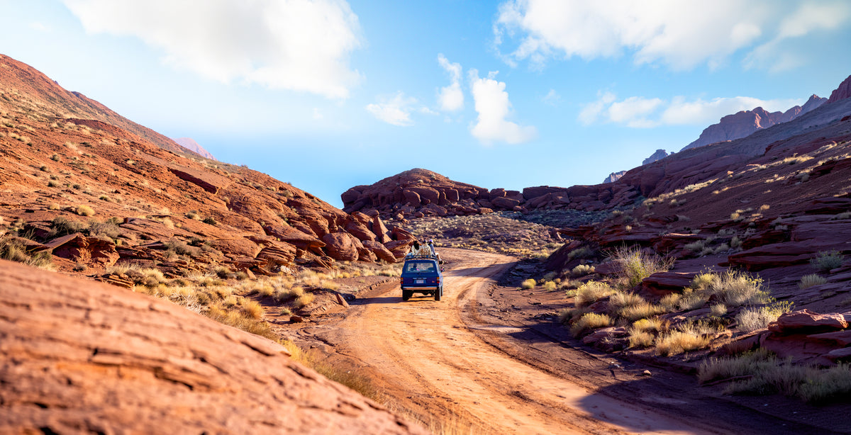 A blue off-road vehicle drives along a dirt trail through a rugged red rock desert canyon under a bright blue sky with scattered clouds.
