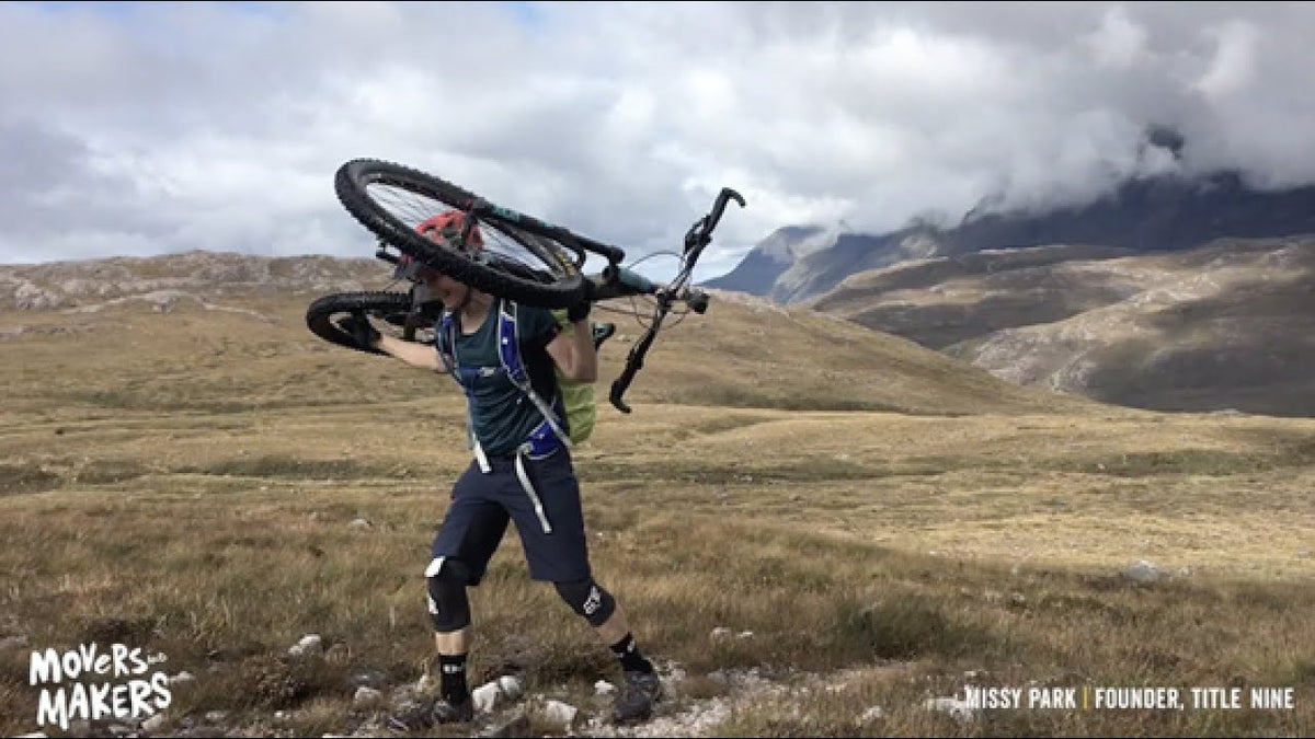 A mountain biker carrying a bike on her back through a grassy mountain landscape under a cloudy sky. Text overlays read 'Movers-Makers' on the bottom left and 'Missy Park | Founder, Title Nine' on the bottom right.