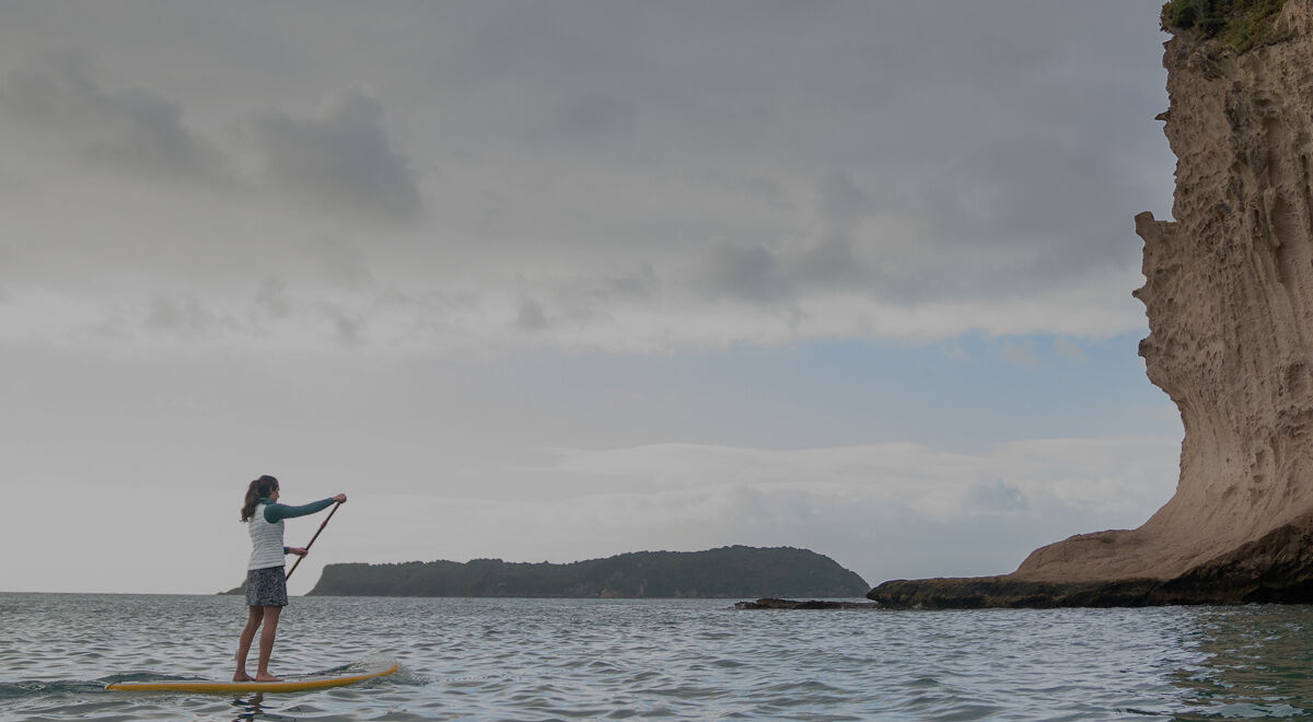 A woman stands on a paddleboard on calm water, holding a paddle near a large rocky cliff under a cloudy sky.