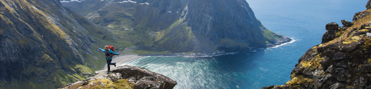 A dramatic mountain and sea landscape with steep rocky cliffs, deep blue water, and a small hiker standing on a cliff edge overlooking the coastline.