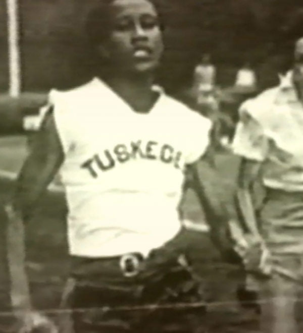 A black and white vintage photograph of a young man wearing a sleeveless athletic jersey with 'TUSKEGA' printed across the chest, standing outdoors.