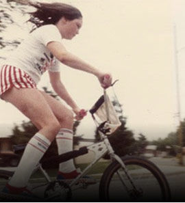 A vintage-style photo of a woman riding a bicycle outdoors, wearing a white top, red-and-white striped shorts, and white knee-high socks.