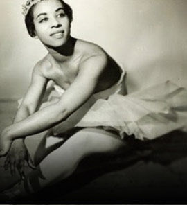 Black and white vintage portrait of a woman seated in a ballet tutu with a tiara, shown against a dark studio background.