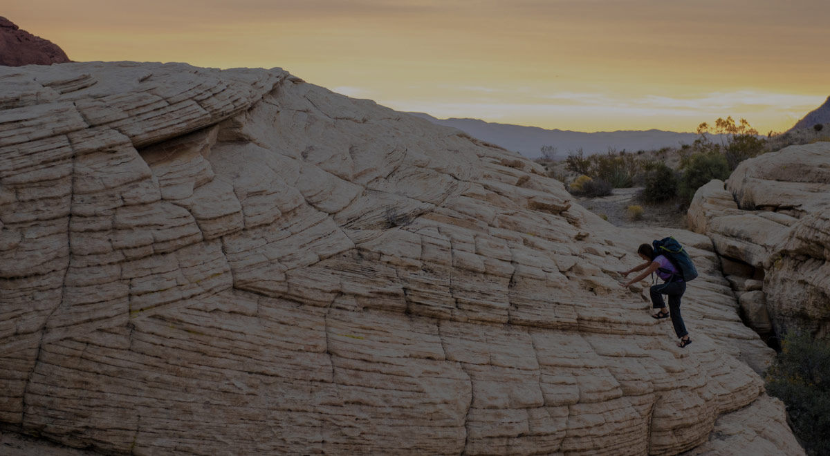 A person hiking or climbing a large layered rock formation in a desert landscape at sunset, with pale sandstone cliffs and distant hills under a warm yellow-orange sky.